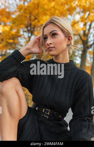 Ein schönes, frisches Mädchen in einem modischen schwarzen Strickkleid sitzt in einem Herbstpark mit goldenem Laub Stockfoto