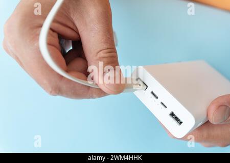 Nahaufnahme einer Hand, die ein USB-Kabel mit einer weißen Powerbank auf blauem Hintergrund verbindet Stockfoto
