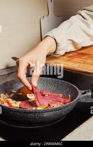 Eine nicht erkennbare Frau fügt dünne Rindfleischscheiben in die Pfanne mit Gemüsedressing Stockfoto