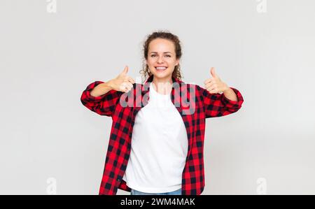 Glückliche, selbstbewusste junge Erwachsene Frau, die Daumen hoch zeigt und vor hellgrauem Hintergrund lächelt. Zufriedener Verbraucher. Stockfoto
