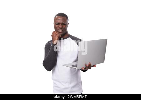 Junger amerikanischer Mann in einem schwarz-weißen Pullover, der einen grauen Laptop auf weißem Hintergrund mit Kopierraum hält Stockfoto