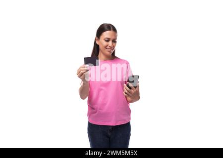 Junge schlanke brünette Frau in rosa Basic-T-Shirt mit Kreditkarte Mockup und Smartphone auf weißem Hintergrund mit Kopierraum Stockfoto