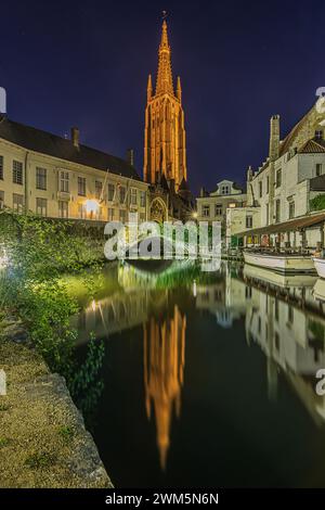 Kanal in der belgischen Hansestadt Brügge am Abend. Kirche unserer Lieben Frau und beleuchtete historische Gebäude zur blauen Stunde. Reflexionen auf Wasser Stockfoto