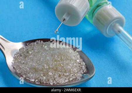 Insulinspritze mit einem Tropfen Insulin in Nahaufnahme am Nadelende und einem Löffel mit Zucker auf blauem Hintergrund. Diabetestherapie, Blutzucker Stockfoto