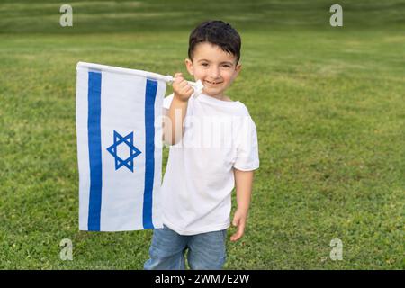 Der lächelnde Junge zeigt stolz die Flagge Israels in einer sonnigen Parklandschaft und drückt Freude und Patriotismus aus. Stockfoto