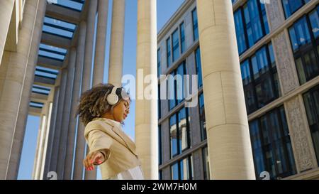 Mit Kopfhörern und einem strahlenden Lächeln tanzt eine junge Berufsfrau frei in einem urbanen Raum, ihre dynamische Bewegung kontrastiert mit den statischen Linien der umgebenden Architektur. Stockfoto