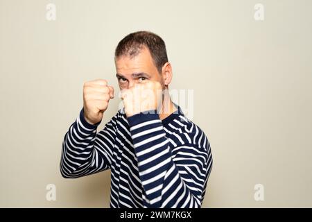 Bärtiger hispanischer Mann in seinen 40ern, der einen gestreiften Pullover trug, mit Fäusten in Verteidigungsposition, bereit zum Kampf, isoliert auf beigefarbenem isoliertem Hintergrund. Stockfoto