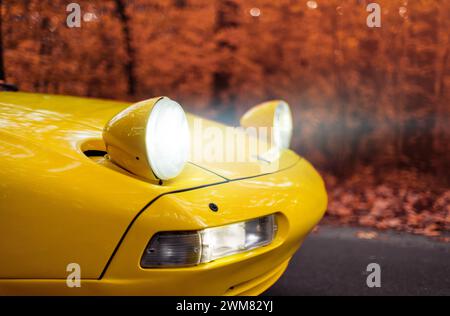 Gelber Porsche 928 im Herbstwald. deutscher Sportwagen der 1980er Jahre im Laubwald. Nahansicht der vorderen Ecke – die einklappbaren Scheinwerfer leuchten hell. Stockfoto