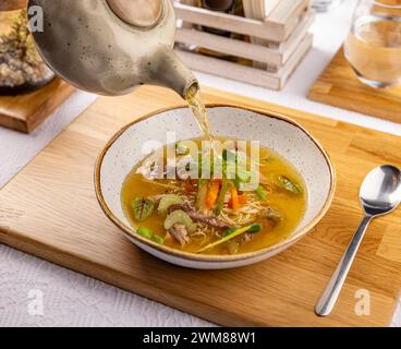 Der Kellner gießt Brühe in eine Hühnersuppe mit Nudeln, Fleisch und Gemüse. Stockfoto
