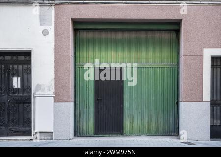 Große grüne Metalltüren einer Garage und eine weitere kleine schwarze Zugangstür im Erdgeschoss mit einem Sicherheitsschloss an der Zement- und Ziegelfassade Stockfoto