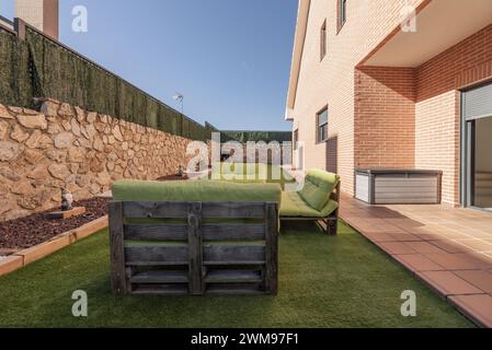 Sitzbereich mit Palettensofas mit grünen Kissen neben einem Backsteinhaus mit Garten und Kunstrasenböden Stockfoto