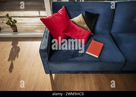 Ein Buch und eine Lesebrille auf einem blauen Samtsofa in einem Zimmer mit Zugang zu einer Terrasse mit Holzfußboden Stockfoto