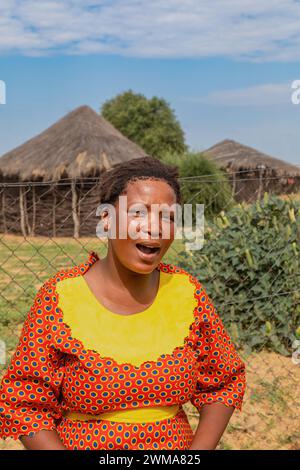 afrikanische Dorffrau, die im Hof steht, in der Hütte im Hintergrund mit Strohdach und blauem Himmel, Südafrika Stockfoto