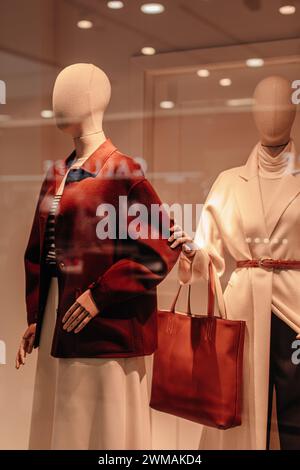 Zwei stilvolle Schaufensterpuppen in einem Schaufenster, gekleidet mit saisonalen Stoffen. Braune Jacke, weißer langer Mantel, braune Ledertasche Stockfoto