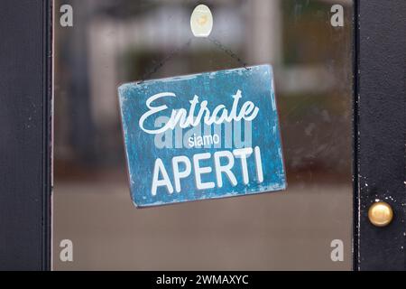 An einer Glastür angebrachtes Schild mit der Aufschrift „Entrate, siamo aperti“, was auf englisch „Kommen Sie rein, wir sind offen“ bedeutet. Stockfoto