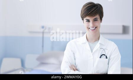 Junge lächelnde Ärztin posiert mit überkreuzten Armen in einem Krankenhauszimmer, medizinische Hilfe und Gesundheitskonzept Stockfoto