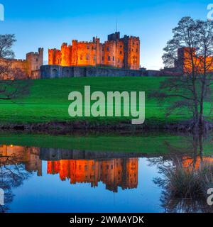 Blick auf Alnwick Castle in Northumberland, beleuchtet bei Nacht, während sie im Fluss ALN von den Weiden aus gesehen reflektiert wird Stockfoto