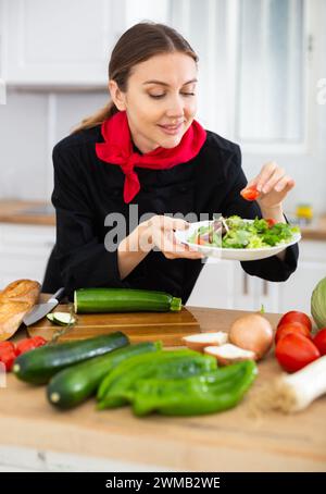 Positive weibliche Küchenchefin in schwarzer Uniform, die Gemüsegericht zubereitet Stockfoto