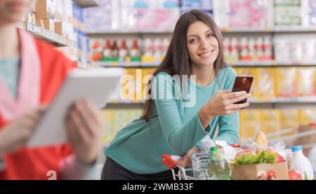 Junge Frau, die ein Smartphone im Supermarkt benutzt und in die Kamera lächelt Stockfoto