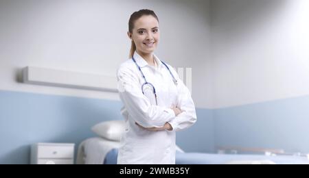 Junge lächelnde Ärztin posiert mit überkreuzten Armen in einem Krankenhauszimmer, medizinische Hilfe und Gesundheitskonzept Stockfoto