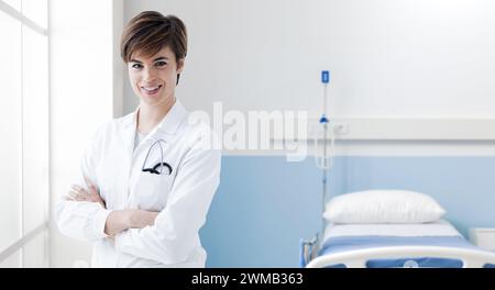 Junge lächelnde Ärztin posiert mit überkreuzten Armen in einem Krankenhauszimmer, medizinische Hilfe und Gesundheitskonzept Stockfoto