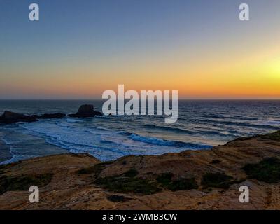 Das Bild zeigt einen wunderschönen Sonnenuntergang über einer zerklüfteten Küste mit Wellen, die gegen die Felsen krachen. Stockfoto