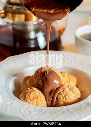 Geschmolzene Schokolade gießt auf Profiterolen. Profiteroles gießen heiße geschmolzene Schokolade. Effuse Schokolade auf Profiteroles auf Restauranttisch Stockfoto