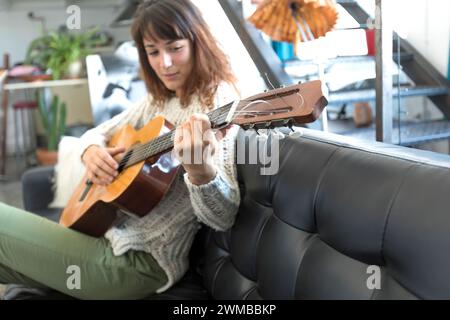 Eine Frau, die in Musik eingetaucht ist, spielt ihre Gitarre, während sie bequem auf einer Ledercouch sitzt. Ihre legere Kleidung und die gemütliche Inneneinrichtung sorgen für einen Intim Stockfoto