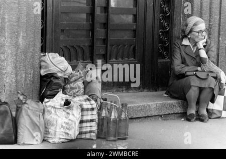 Bukarest, Rumänien, Februar 1990. Kurz nach der antikommunistischen Revolution im Dezember 1989 bleiben die Menschen immer noch lange in der Schlange, um sich an grundlegende Lebensmittel zu halten. Das zentralisierte sozialistische Wirtschaftssystem schuf Knappheit und Hunger. Hier sitzt eine ältere Frau und wartet in der Schlange auf die Lieferung von Milch. Andere Leute ließen ihre Taschen mit wiederverwendbaren Glasflaschen zurück und reservierten ihren Platz in der Schlange. Stockfoto