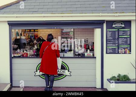 Frau in rotem Mantel, die an einem Speise- und Getränkekiosk an der Promenade in Teignmouth, South Devon, am Meer serviert wird. Stockfoto