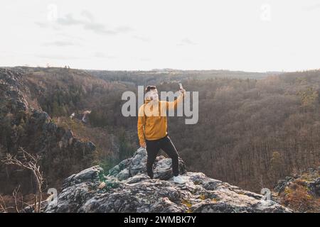 Ein Reisender in einer gelben Jacke, der am Rand einer Klippe steht und bei Sonnenuntergang ein Selfie auf seinem Handy macht. Divoka-Sarka-Tal, Prag. Stockfoto