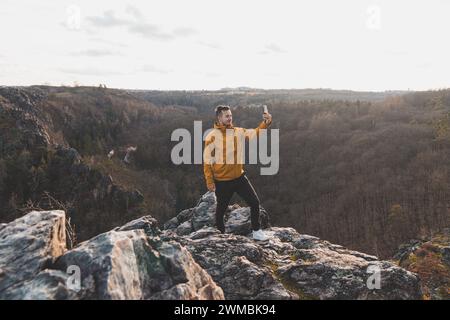 Ein Reisender in einer gelben Jacke, der am Rand einer Klippe steht und bei Sonnenuntergang ein Selfie auf seinem Handy macht. Divoka-Sarka-Tal, Prag. Stockfoto