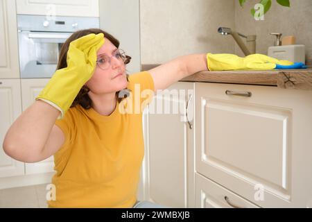 Eine müde Frau nach dem Putzen hält sich an einem schmerzenden Kopf in einer Küche fest, Handarbeit. Traurige Frau in gelben Handschuhen beim Putzen des Hauses Stockfoto