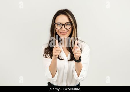 Fröhliche Frau mit einer Brille, die die Daumen hoch hält, eine weiße Bluse und Jeans trägt Stockfoto