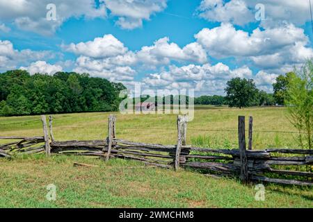 Ein handgebauter rustikaler Holzzaun mit Stacheldraht rund um das Ackerland mit einer alten Scheune im Hintergrund auf der Weide an einem sonnigen Tag mit weißem flauschigem Wasser Stockfoto