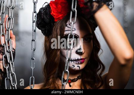 Eine Frau mit auffälligem Schädel-Make-up schaut durch hängende Ketten, ihre Augen erzählen eine Geschichte von Mystik. Der rot-schwarze Kopfschmuck mit Blumenmuster steht im Kontrast zum industriellen Ambiente. Stockfoto