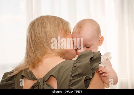 Aus nächster Nähe hält die junge Mutter ihr Baby in den Armen und küsst sie, an einem sonnigen Tag zu Hause, sanft fokussiert Stockfoto