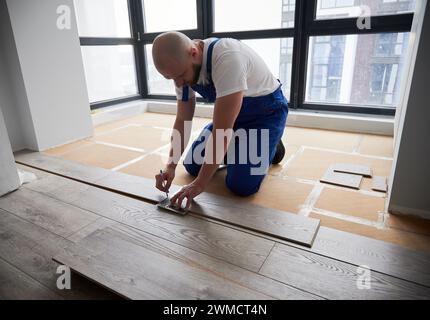 Mann in Arbeitsanzug mit Metalllineal und Stift beim Zeichnen von Linien auf Laminatfußböden. Männlicher Fußbodenmonteur bereitet Laminatplanke für die Installation in einer Wohnung vor, die gerade renoviert wird. Stockfoto