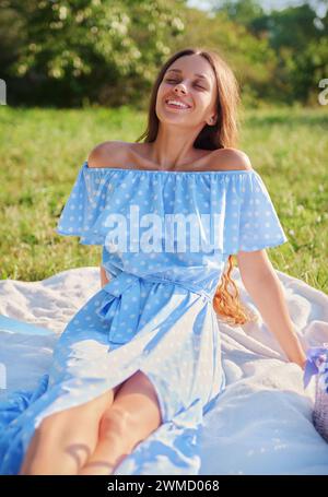 Picknickszene: Glückliches lächelndes Mädchen, das in einem Park auf einem Karo sitzt und sich sonnt. Außenporträt einer schönen fröhlichen jungen Frau in blauem Kleid Stockfoto