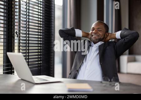 Ein zufriedener afroamerikanischer Unternehmer in einem stilvollen Anzug macht mit seinem Laptop eine friedliche Pause an seinem Arbeitsplatz. Stockfoto