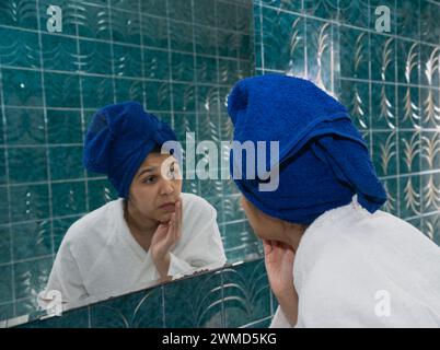 Junge Frau im Badezimmer, die in den Spiegel blickt, mit Bademantel und Tuch auf dem Kopf Stockfoto