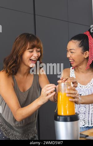 Junge kaukasische Frau und junge birassische Frau lachen zusammen in einer Küche und machen Smoothie Stockfoto