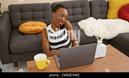 Fesselnde afroamerikanische Frau, die sich am Telefon lebhaft unterhalten, wunderschön auf dem Sofa zuhause, selbstbewusst auf der Arbeit auf dem Laptop jongliert und gleichzeitig genießt Stockfoto