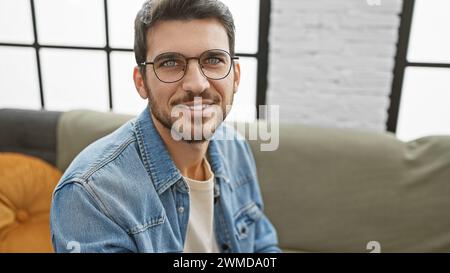 Hübscher junger hispanischer Mann mit Brille und Bart, der in einem stilvollen Wohnzimmer lächelt. Stockfoto