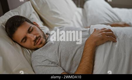Ein junger hispanischer Mann, der sich friedlich auf einer weißen Bettdecke in einem gemütlichen Schlafzimmer ausruhen lässt. Stockfoto