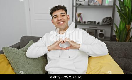 Ein fröhlicher junger Mann macht mit seinen Händen eine Herzform, während er auf einem Sofa in einem gemütlichen Wohnzimmer sitzt. Stockfoto