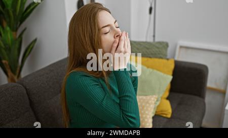 Eine müde junge Frau, die drinnen gähnt, während sie auf einer Couch sitzt und den natürlichen Lebensstil im Wohnzimmer darstellt. Stockfoto