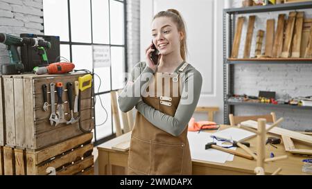 Eine junge blonde Frau in einer Tischlerei, die am Telefon telefoniert, während sie drinnen lächelt. Stockfoto