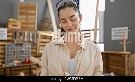 Schöne junge hispanische Frau, ein lächelnder Zimmermann, der stolz in ihrer Tischlerei steht und eine Schutzbrille trägt Stockfoto