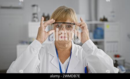 Engagierte, blonde Wissenschaftlerin mittleren Alters, in Brille gekleidet, mit ernstem Ausdruck, während sie fleißig am Labortisch arbeitet, vertieft in Medi Stockfoto